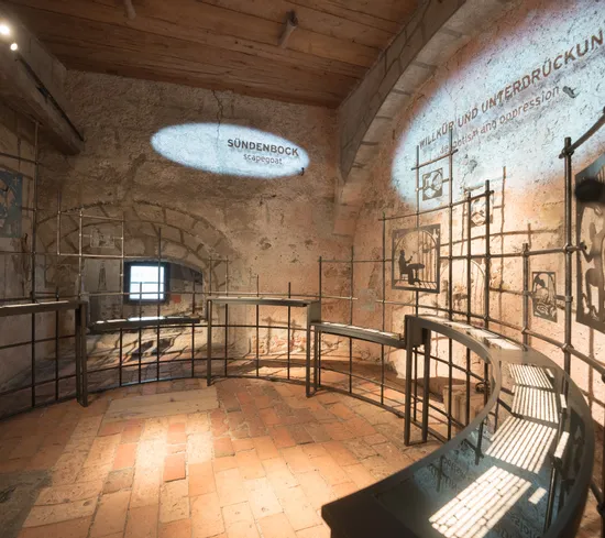 Historische Museumsausstellung über Sündenböcke und Unterdrückung in einem Raum mit Steinwänden und Metallrahmen